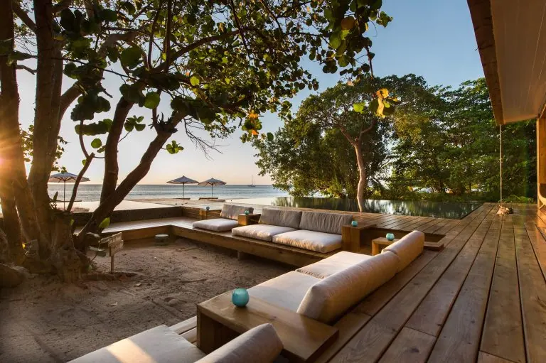A waterfront lounge area in Roatan, Honduras, features wooden decks, comfortable beige seating, and a view of the ocean with umbrellas and sailboats in the distance, surrounded by lush green trees at sunset.