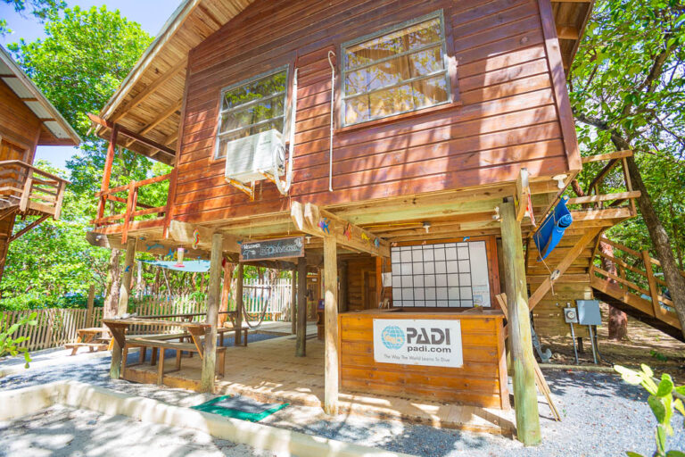 A two-story wooden building in Roatan, Honduras, with an air conditioning unit on the upper floor and a PADI dive shop sign on the lower level, surrounded by lush green trees and outdoor seating areas.