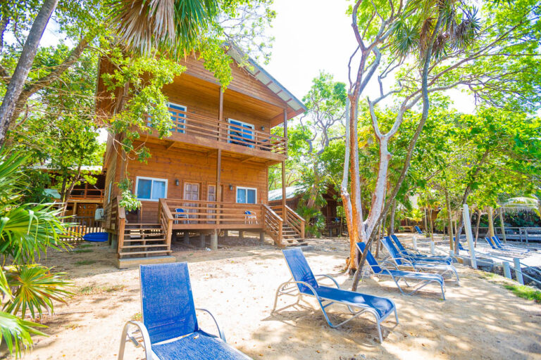 A wooden two-story beach house nestled among lush green trees on Roatan, Bay Islands, Honduras, with a sandy area and blue lounge chairs in the foreground.