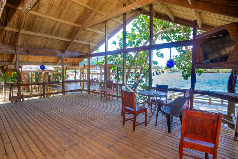 A rustic wooden pavilion with a thatched roof overlooking Roatan's waterfront in Honduras, featuring a small seating area with chairs and a table, surrounded by lush greenery and trees.