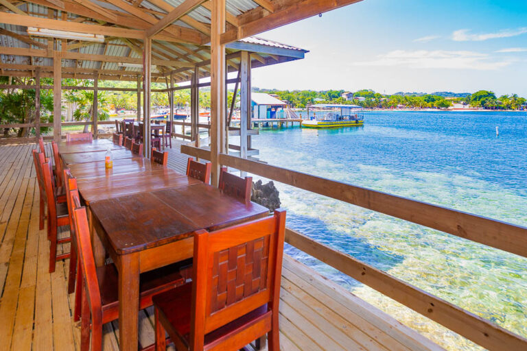 A wooden outdoor dining area on Roatan with a long table and chairs overlooking clear blue Caribbean waters and the Honduran coastline.
