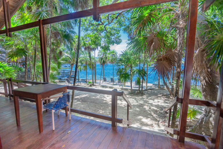 A wooden deck with a table and a woven chair overlooks a tropical beach in Roatan, Honduras, with lush palm trees and the blue Caribbean Sea in the background.