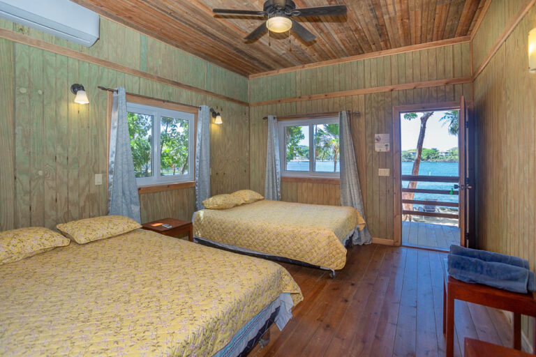 Two double beds with yellow floral bedding in a wooden-paneled hotel room on Roatan Honduran Bay Islands, featuring windows with ocean views and a door leading to a balcony overlooking the water.