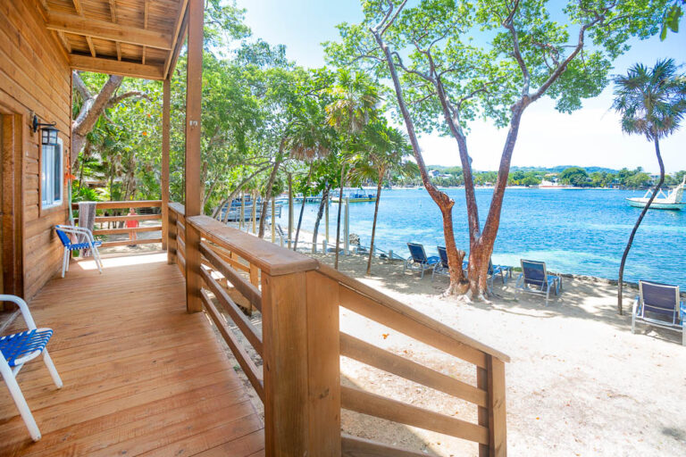 Wooden balcony overlooking a sandy beach with lounge chairs, lush green trees, and clear blue waters in Roatan, Honduras, with boats anchored offshore.