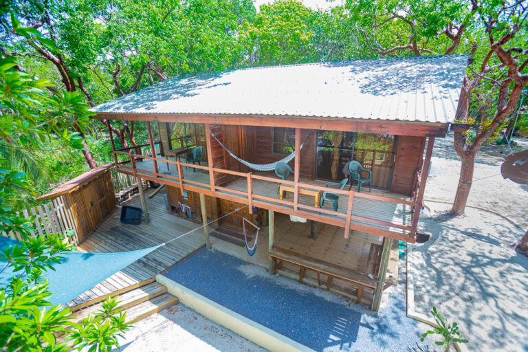 A two-story wooden vacation cottage with a metal roof located on Roatan in the Bay Islands of Honduras, surrounded by lush green trees with a spacious outdoor deck featuring chairs, a hammock, and a shaded area with a slanted roof.