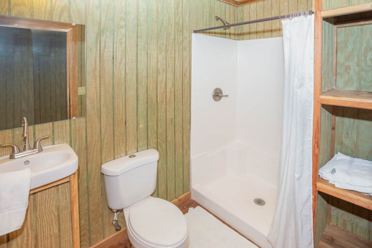 A compact bathroom in Roatan, Honduras, featuring a wood-paneled wall, a white toilet, a small white sink with a silver faucet, a mirror above the sink, a shower with a white curtain, and wooden shelves holding folded towels.