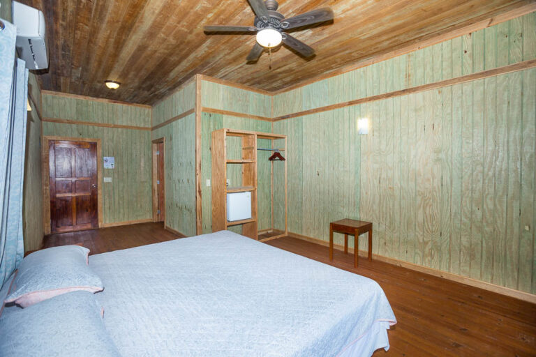 A cozy bedroom in a Roatan hotel or guesthouse featuring green wooden panel walls, a wooden ceiling with a ceiling fan, a bed with white bedding, a small wooden stool, and a partial view of a door leading to another room or hallway in Honduras.