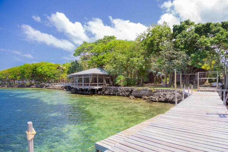 A wooden pier extends over clear turquoise waters towards a small gazebo surrounded by lush green trees on Roatan, Honduras, under a bright blue sky with scattered clouds.