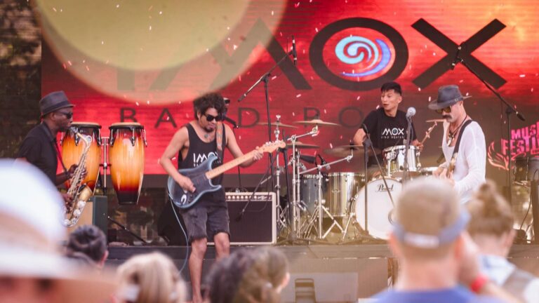 A live band performs on an outdoor stage in Roatan, Honduras, with musicians playing guitars, drums, percussion, and saxophone during a festival or event.