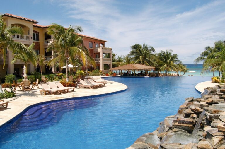 A luxurious resort with a large infinity pool, surrounded by palm trees and a view of the ocean on Roatan, Honduras, with a thatched-roof bar area and multi-story buildings with balconies.