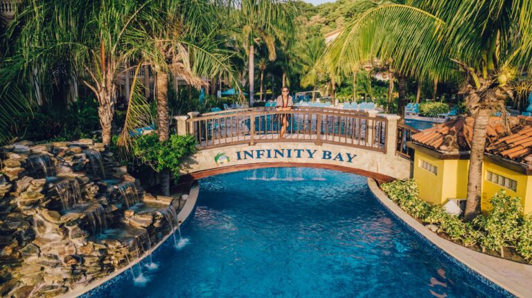 A woman stands on a wooden bridge over a swimming pool at Infinity Bay resort, surrounded by lush palm trees and tropical landscaping in Roatan, Honduras.