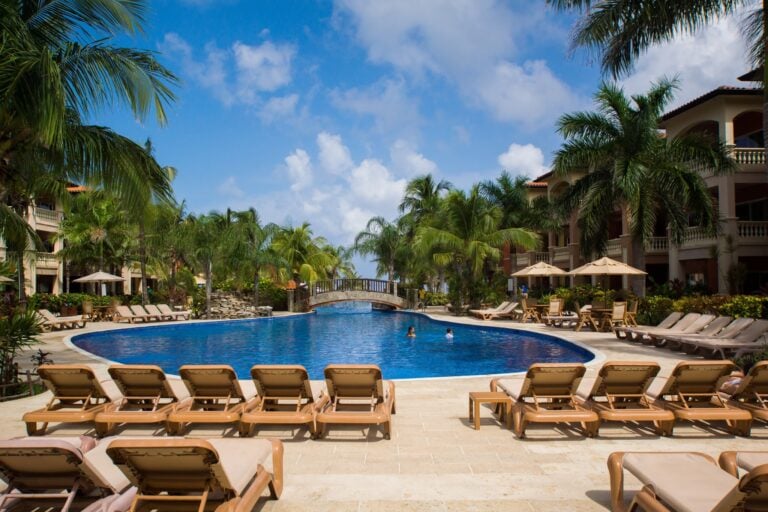 A resort pool area on Roatan, Honduras, features lounge chairs, a large swimming pool, palm trees, tropical landscaping, and a building with balconies under a partly cloudy blue sky.