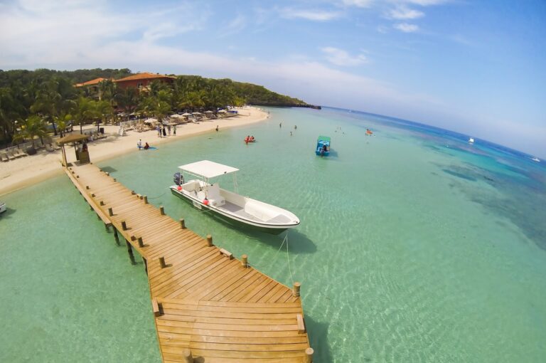 A wooden pier extends into clear turquoise waters near a sandy beach lined with palm trees and beach chairs at a resort in Roatan, Honduras, with boats and people enjoying the water.