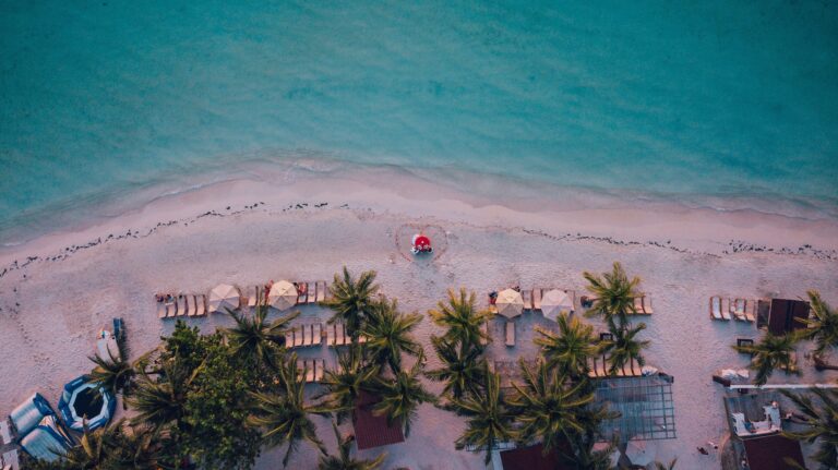 An aerial view of a sandy beach with palm trees, lounge chairs, umbrellas, and a heart-shaped decoration on the shore, overlooking the turquoise waters of Roatan, Honduras.