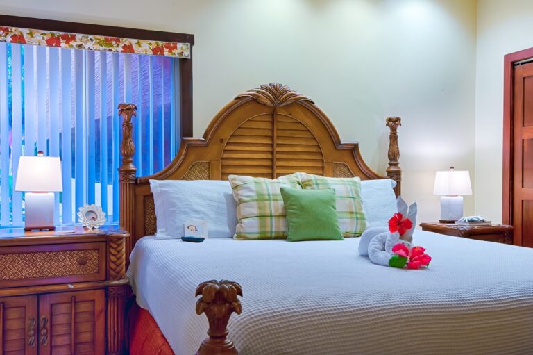 A bedroom with a wooden headboard, white bedding with green plaid pillows, and a towel shaped like a swan with hibiscus flowers on it, set in a hotel on Roatan, Honduras.