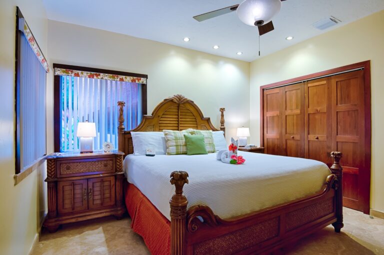 A hotel room in Roatan, Honduras features a large wooden bed with tropical-themed towels folded into swans and flower decorations, a wardrobe, bedside tables with lamps, and a window with vertical blinds and floral valances.