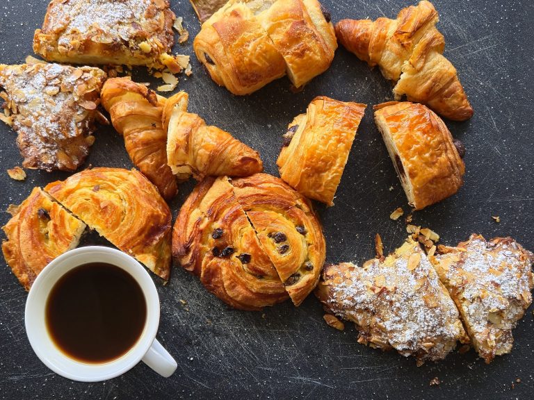 A variety of freshly baked pastries, including croissants and danishes, are displayed on a black surface with a cup of coffee underneath, with a small container of syrup in front, on Roatan, Honduras.