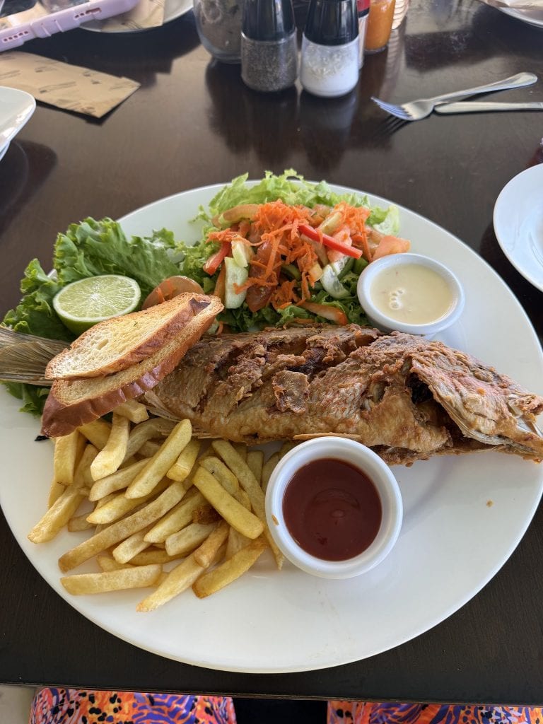 A plate of Jamaican fried fish with French fries, fresh salad with shredded carrots and lettuce, a lime wedge, garlic bread, and dipping sauces, set on a table in Roatan, Honduras.