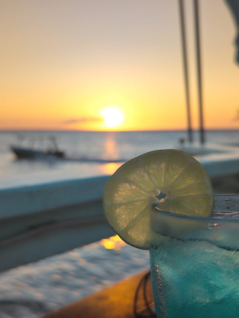 A slice of lemon and a blue frozen drink garnished with a lemon wedge are placed on a table aboard a boat, with a sunset over the ocean and sailboat masts visible in the background in Roatan, Honduras.