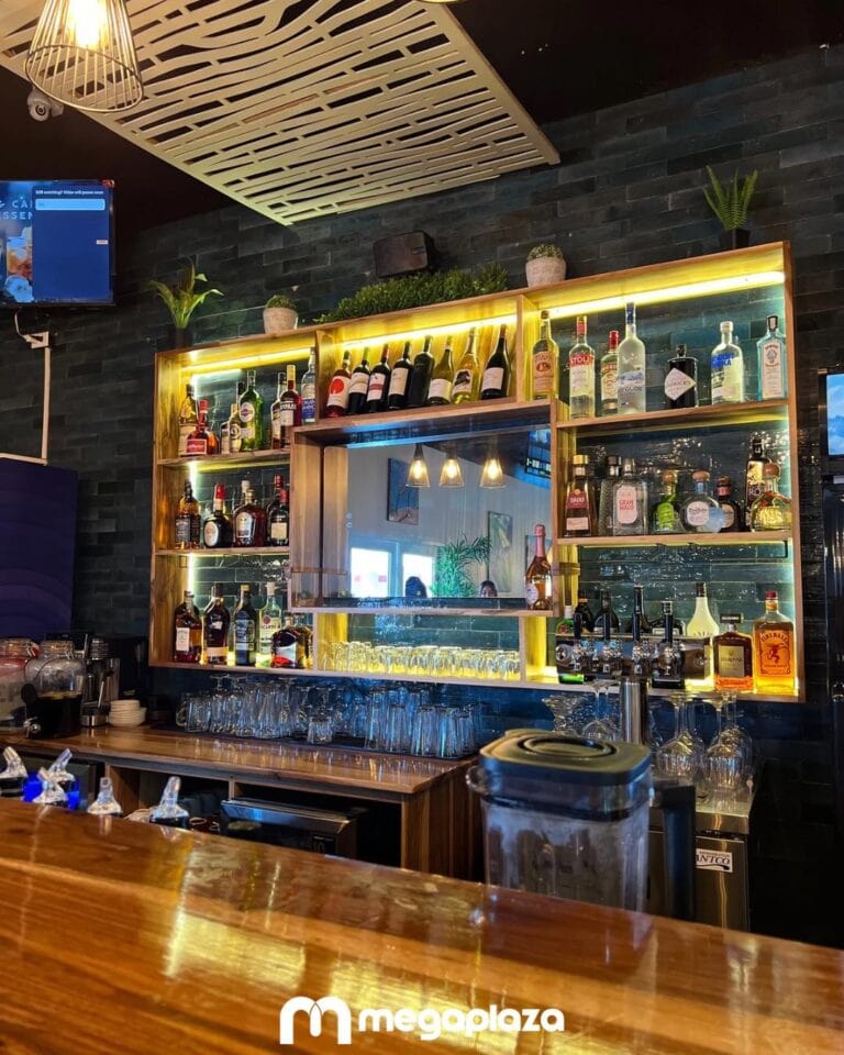 A well-lit bar area in Roatan, Honduras, features a wooden and glass liquor shelf displaying various bottles of spirits, along with glassware, potted plants on top, and a mirror reflecting the interior, all set against a dark wall with a decorative ceiling panel.