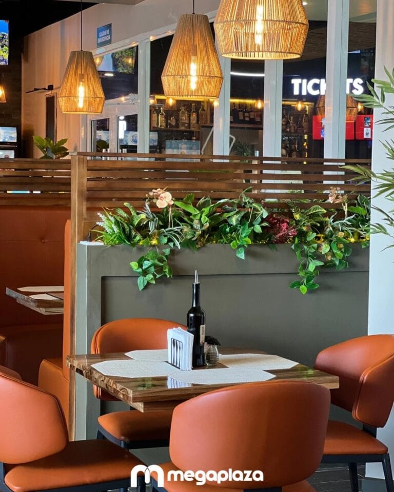 A cozy restaurant interior in Roatan, Honduras, features a wooden table with orange chairs, a decorative planter with lush green plants and flowers, pendant lights with woven shades, and a view of an open bar area through large glass windows.