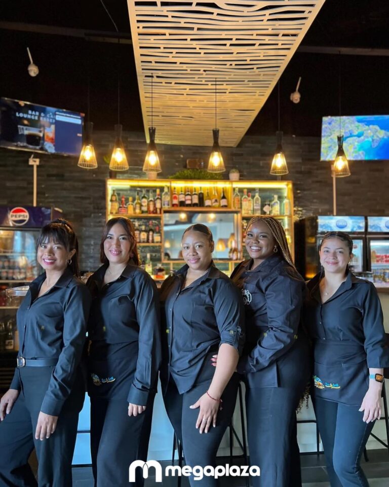 A group of five smiling female staff members dressed in black uniforms stand inside a bar or restaurant in Roatan, Honduras, with a backdrop of liquor bottles, a bar counter, and warm lighting fixtures.