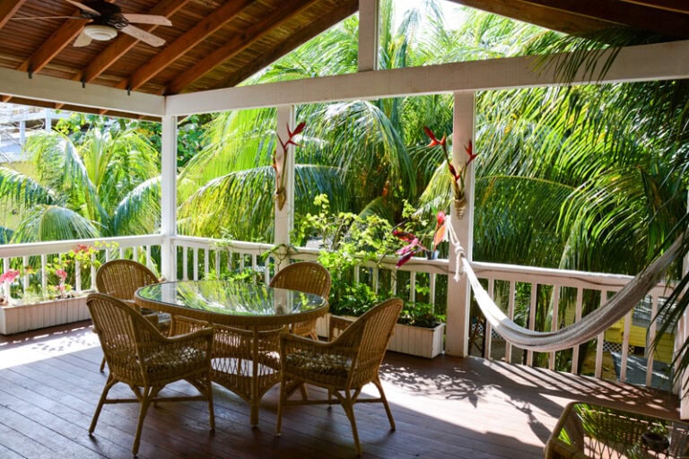 A shaded outdoor porch in Roatan, Honduras, features a glass-topped dining table surrounded by wicker chairs, with lush tropical greenery and palm trees visible beyond the white railing, and a hammock hanging from the ceiling beams.