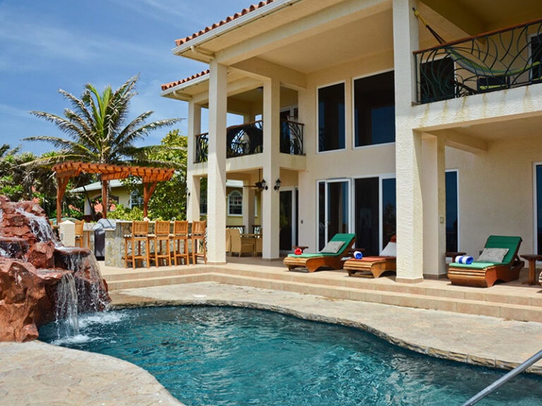 A luxury villa in Roatan, Honduras, features a swimming pool with a waterfall, multiple lounge chairs with towels on a patio, a covered outdoor dining area with a wooden table and chairs, and a two-story building with large windows and balconies surrounded by lush tropical greenery.