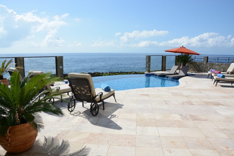 A luxurious swimming pool area with lounge chairs, an orange umbrella, and tropical plants overlooking the ocean on Roatan, Honduras, with a clear blue sky and fluffy clouds.