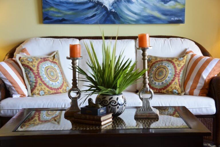 A cozy living room in Roatan, Honduras, features a white sofa with colorful patterned pillows, a glass-top coffee table with stacked books, a decorative black-and-white ceramic vase with green plants, two orange candles on silver candleholders, and a vibrant abstract painting of ocean waves hanging on a yellow wall.