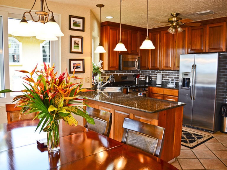 A modern kitchen with wooden cabinets, stainless steel appliances, a granite countertop island, and pendant lighting, set in a cozy home on Roatan, Honduras, with a dining table featuring a colorful flower arrangement, decorative wall art, and a bright window illuminating the space.
