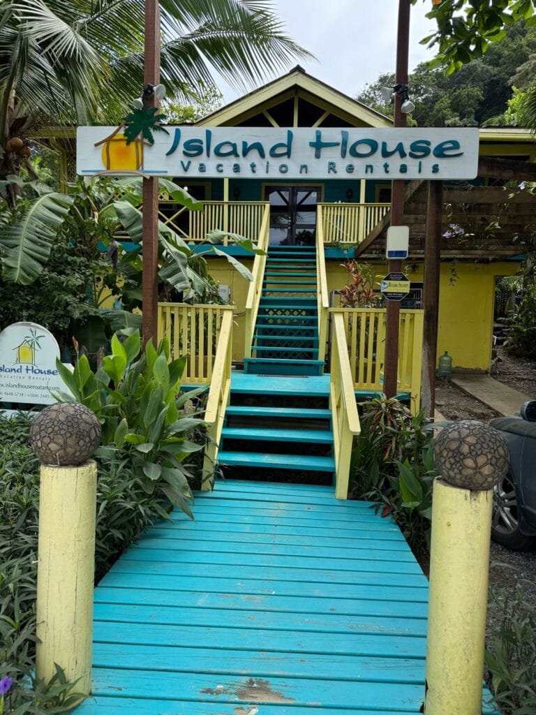 A brightly painted yellow and green two-story vacation rental with a turquoise staircase leading to a front porch, surrounded by lush tropical plants and trees, with signs indicating "Island House Vacation Rentals" in Roatan, Honduras.