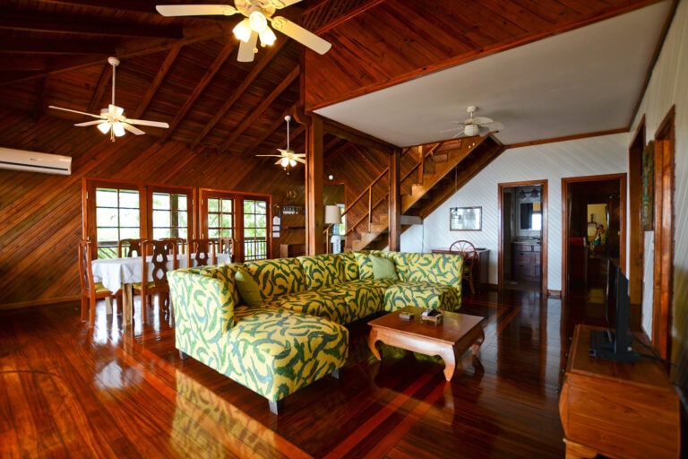 A spacious, open-concept living area in a Roatan, Honduras home features a high wooden vaulted ceiling with ceiling fans, a colorful patterned sofa, a wooden dining table with chairs, and a staircase leading upstairs, all with polished hardwood floors and wood-paneled walls.