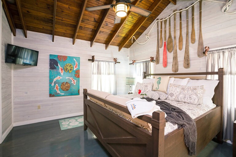 A cozy bedroom in Roatan, Honduras features white wooden walls, a vaulted wooden ceiling, a large bed with decorative pillows and a blanket, hanging spoons as wall decor, and windows with white curtains.