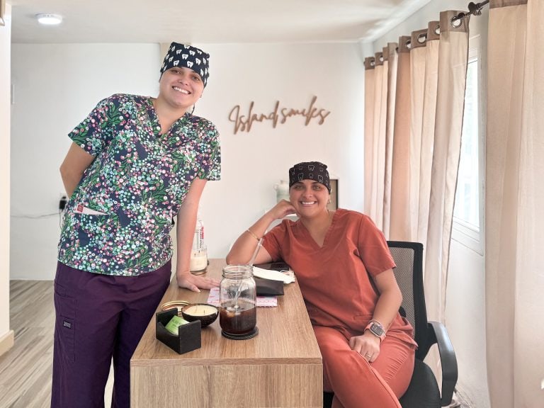 Two women in medical scrubs and bandanas smile inside a restaurant or café in Roatan, Honduras, with beige curtains, a white wall, and a wooden sign reading "Island Smiles" in the background.