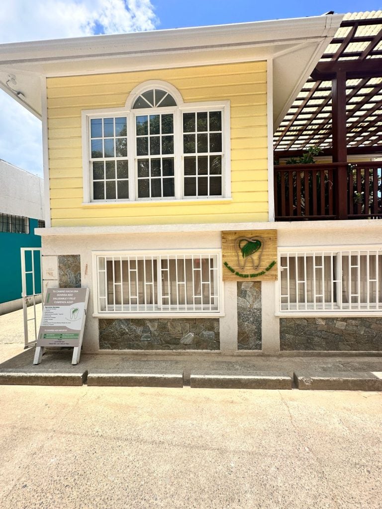 The yellow and white two-story building in Roatan, Honduras, features large windows, a dental clinic sign with a tooth and palm leaf logo, and a sidewalk sign promoting dental health services outside the entrance.