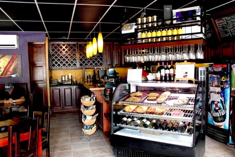 A cozy café in Roatan, Honduras, features a wooden counter with a glass display case of baked goods, hanging wine glasses above, a row of yellow candles on a shelf, and seating with a customer wearing a cap, all set against a dark ceiling and warm-toned walls.