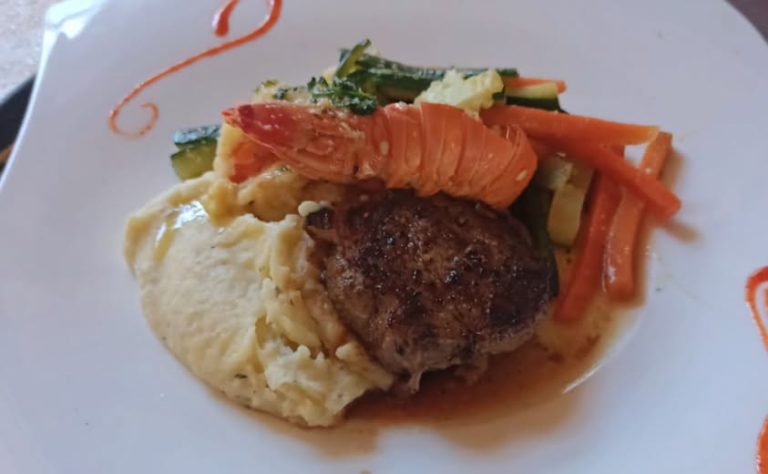 A plate of mashed potatoes, a grilled meat patty, a cooked lobster tail, and sautéed mixed vegetables including carrots and zucchini is shown on a white dish with orange decorative accents, set against a background suggesting a dining setting in Roatan, Honduras.
