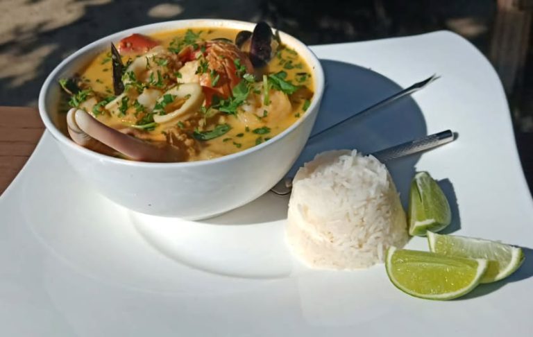 A bowl of flavorful seafood coconut curry garnished with fresh herbs is served with a side of white rice and lime wedges on a white plate, set outdoors in Roatan, Honduras.