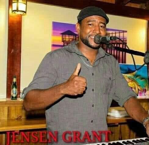 A man in a gray shirt and black cap performs with a microphone and keyboard in a cozy indoor setting in Roatan, Honduras, with colorful artwork and a small bar area visible behind him.