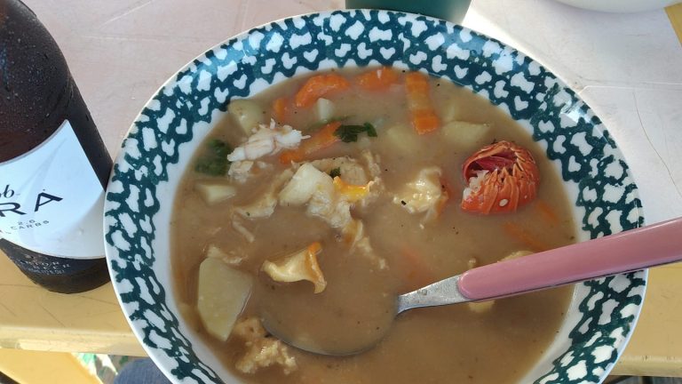 A bowl of hearty chicken, vegetable, and potato soup with a pink-handled spoon on a yellow table, accompanied by a bottle of local Honduran soda, is displayed in a business listing for Roatan, Honduras.