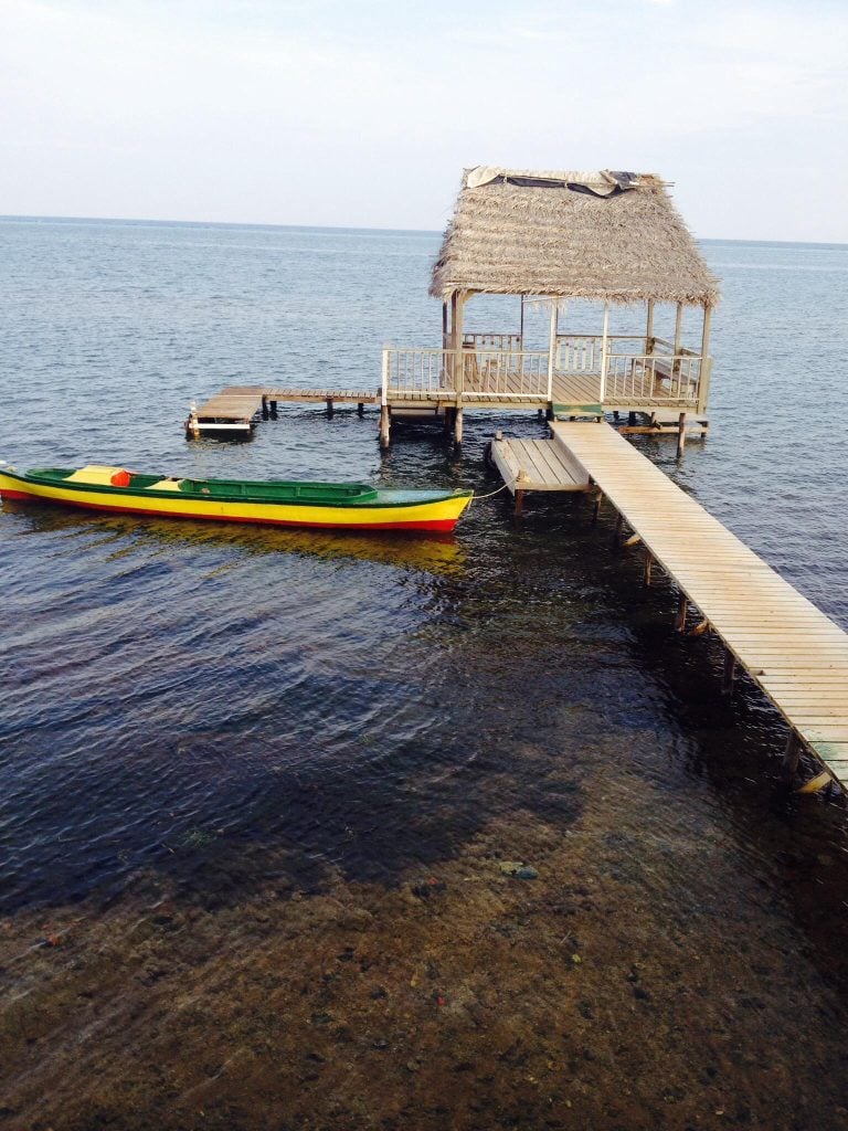 A thatched-roof cabana extends over the clear waters of Roatan, Honduras, connected by a wooden pier with a colorful canoe floating nearby in this coastal scene.