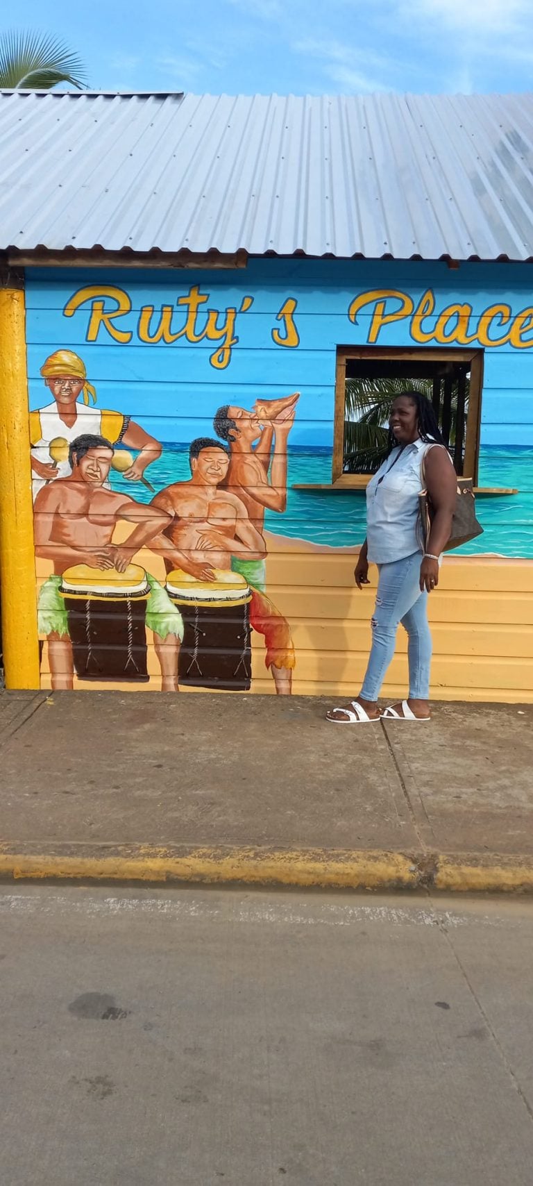A woman stands in front of a colorful mural on a building in Roatan, Honduras, depicting musicians playing drums and a person drinking from a coconut, with the text "Ruty's Place" painted above.