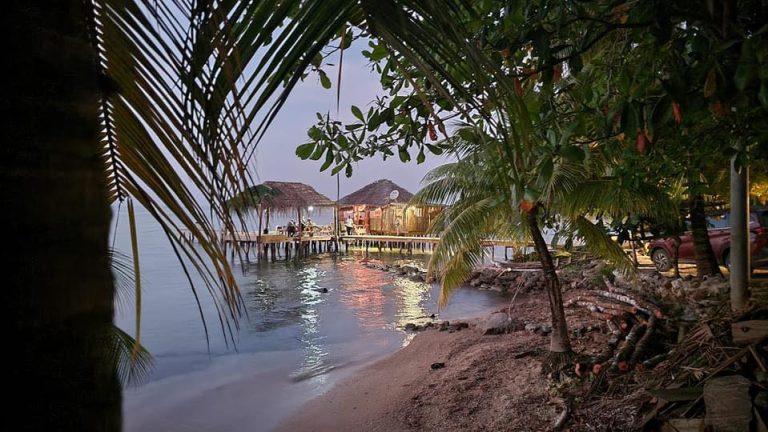 A view of a beachfront restaurant with thatched-roof huts and outdoor seating on a wooden deck, surrounded by lush tropical trees and foliage, on the coast of Roatan, Honduras.
