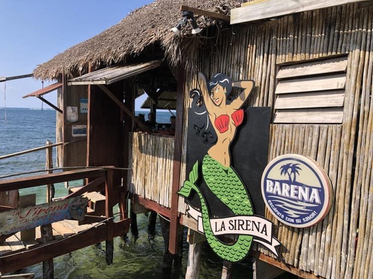 The image shows a wooden overwater structure with a thatched roof in Roatan, Honduras, featuring a colorful sign of a mermaid with long black hair, a red seashell bikini top, and a green scaly tail, along with a circular logo reading "BARENA" and "LA SIRENA.