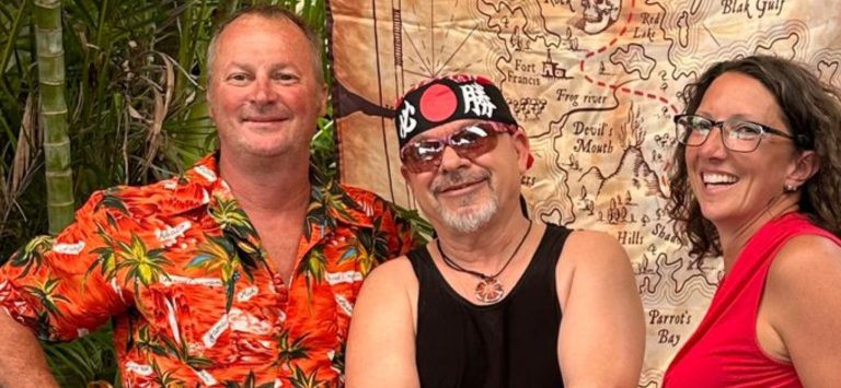 Three smiling people stand in front of a map of Roatan, Honduras, decorated with tropical plants and vibrant attire, indicating a relaxed social setting on the Bay Islands.
