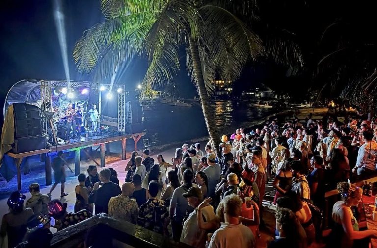 A large crowd gathers on the beach in Roatan, Honduras, to enjoy a live music performance on an elevated stage illuminated by colorful lights, with palm trees and boats visible near the water at night.
