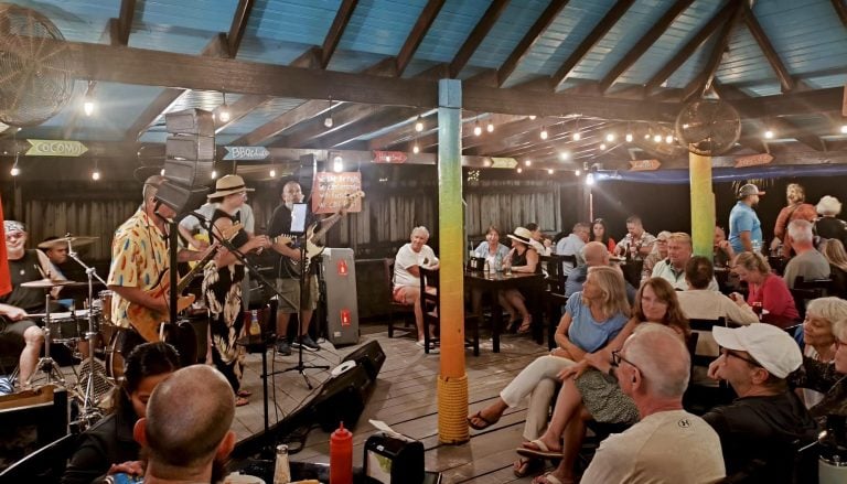 A lively indoor scene in Roatan, Honduras, features a live band performing music to an audience seated at tables under a wooden ceiling with string lights and colorful signs, creating a festive atmosphere.