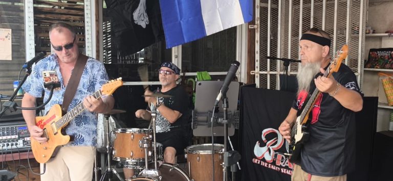 A live band performing with guitar and drums at an outdoor venue in Roatan, Honduras, with a blue and white flag hanging in the background.