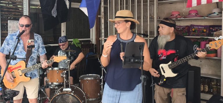 A live band performs on a covered outdoor stage in Roatan, Honduras, with musicians playing guitar, drums, and bass while a woman in a hat and glasses sings into a microphone.